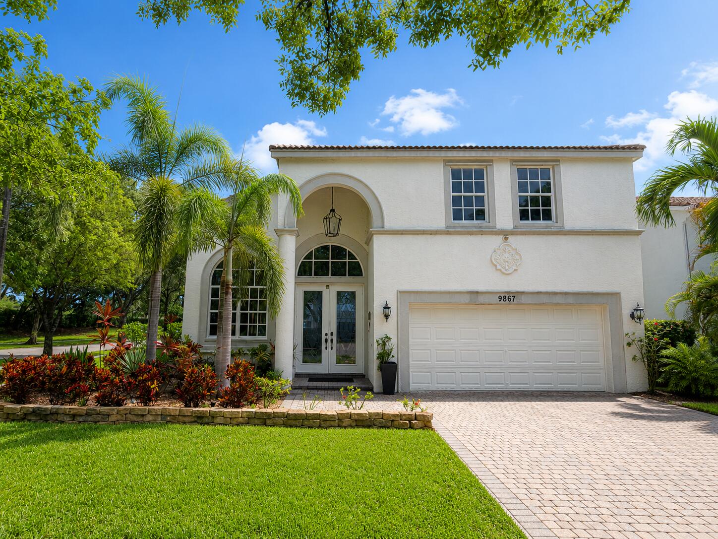 Stucco exterior featuring an arched entryway with double doors, symmetrical windows, a paver driveway, and a manicured lawn with tropical landscaping