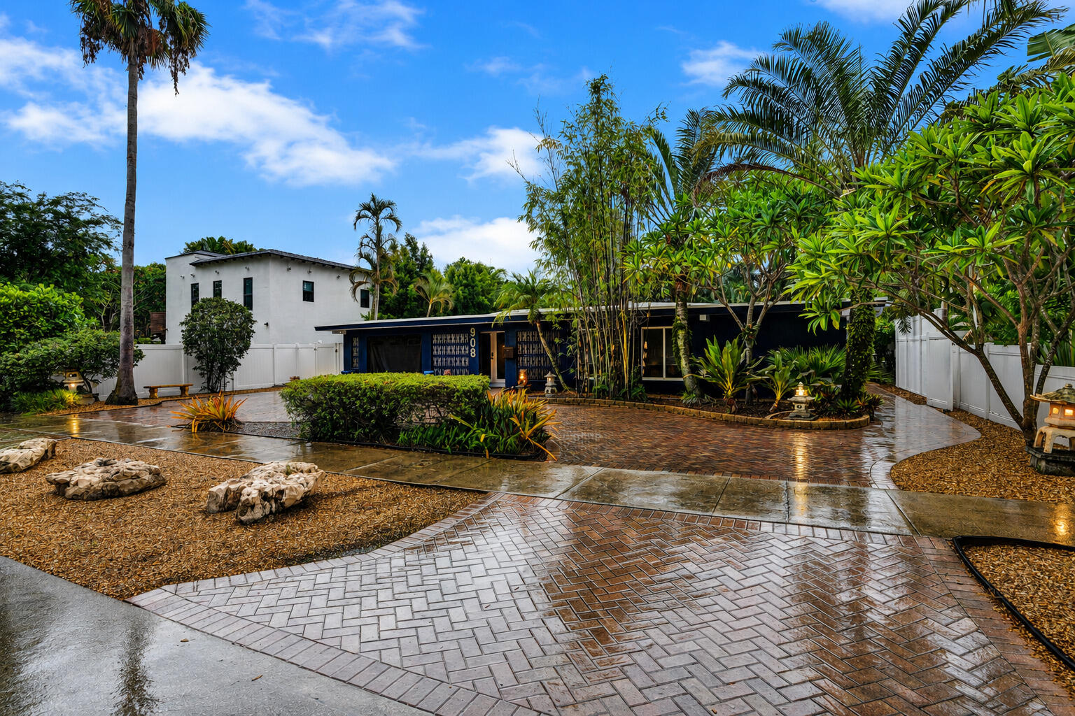 Paved driveway featuring a herringbone pattern, gravel landscaping with natural rock elements, mature palm trees, and lush tropical foliage