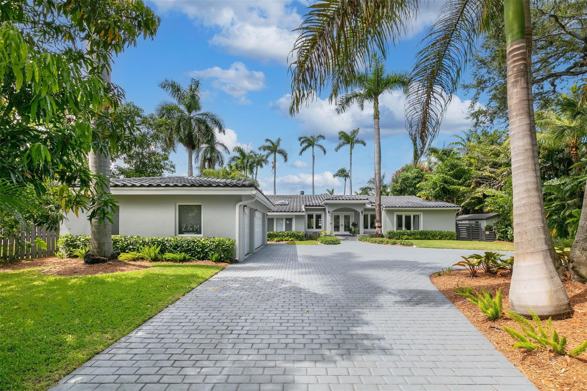 Contemporary residence featuring a paver driveway, lush tropical landscaping, mature palm trees, and a tiled roof