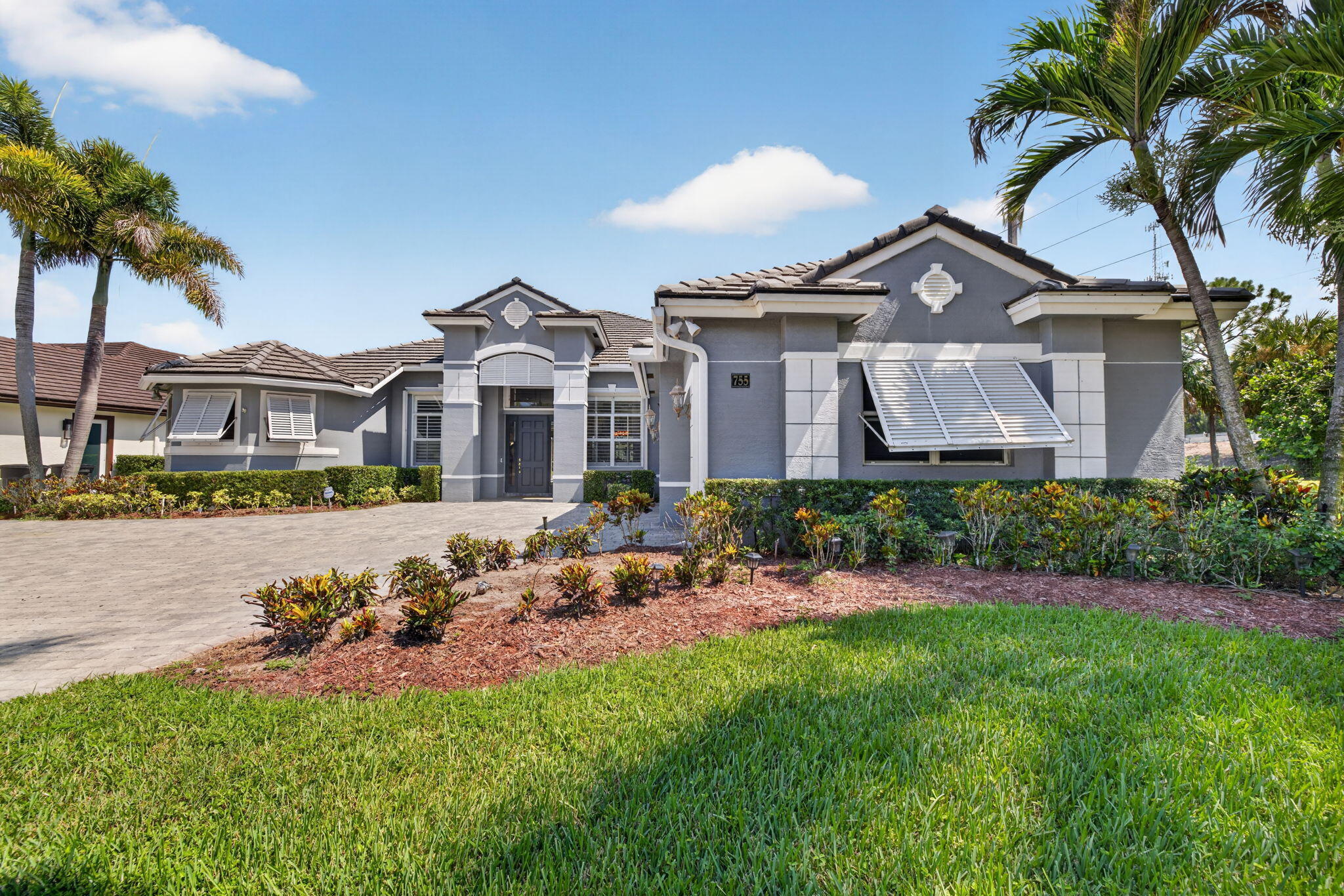Modern home featuring a sophisticated gray and white exterior with architectural detail work, solar panel installation on the roof, mature palm tree landscaping, and a professionally manicured front lawn with colorful mulched planting beds and established shrub borders