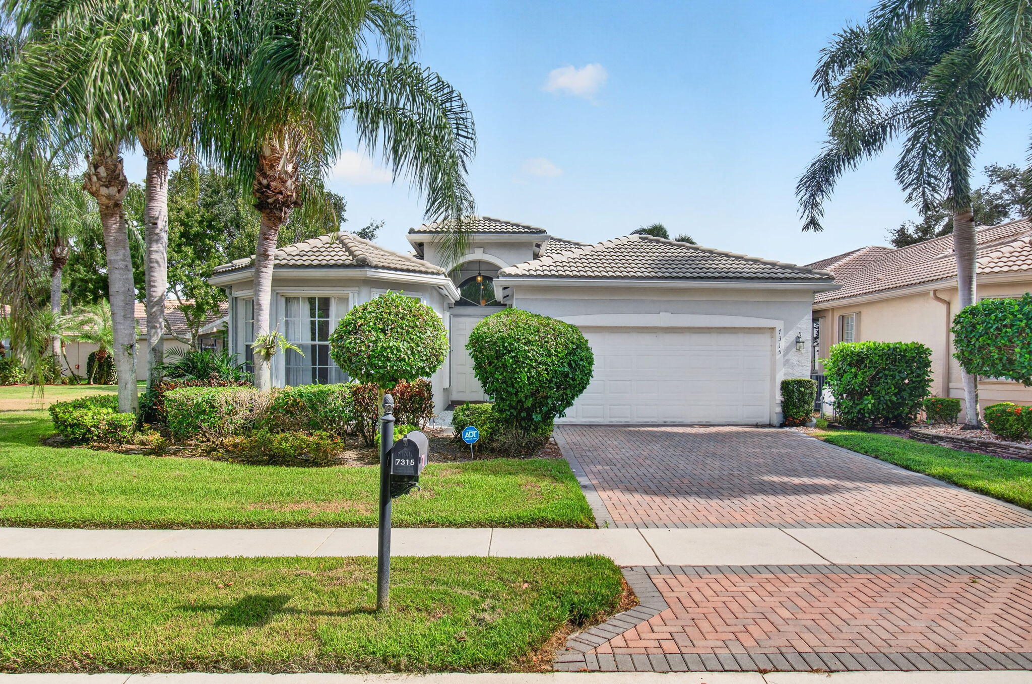 Stucco exterior with a tiled roof, mature palm trees, and a brick paver driveway