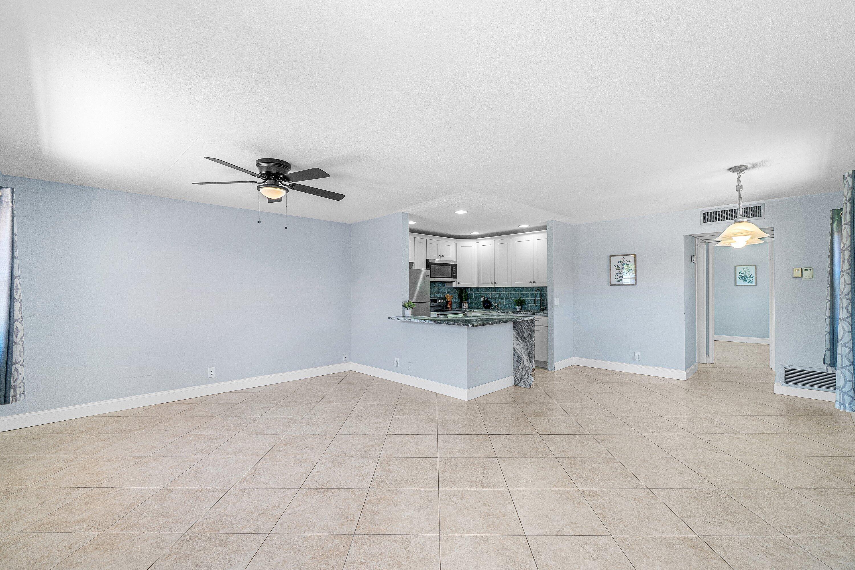 Spacious room with large format tile flooring, a ceiling fan, and a kitchen featuring white cabinetry, a tiled backsplash, stainless steel appliances, and a peninsula with a stone countertop