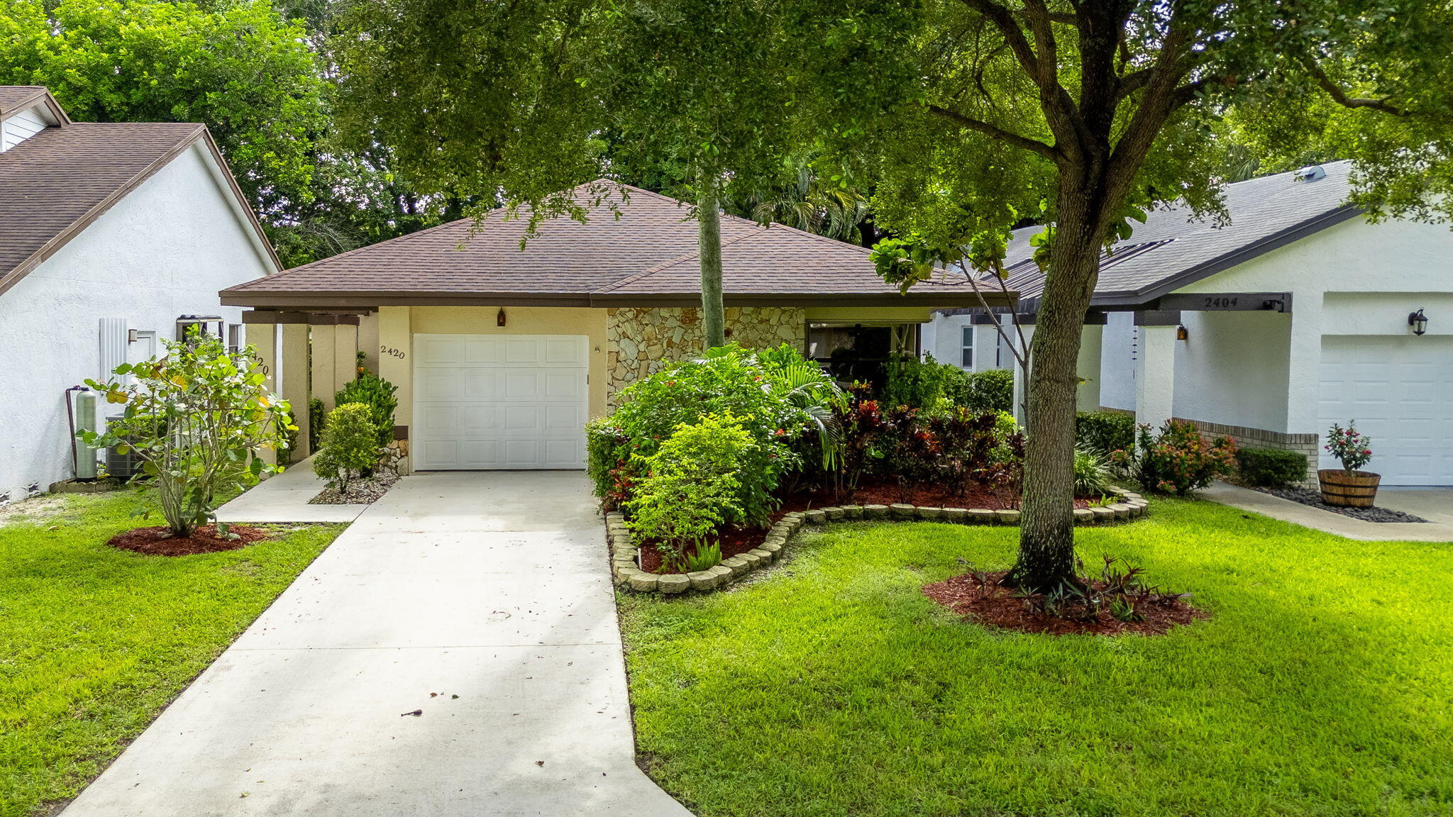 Single-story residence featuring a paved driveway, attached garage, stone veneer facade, mature trees, and landscaped grounds