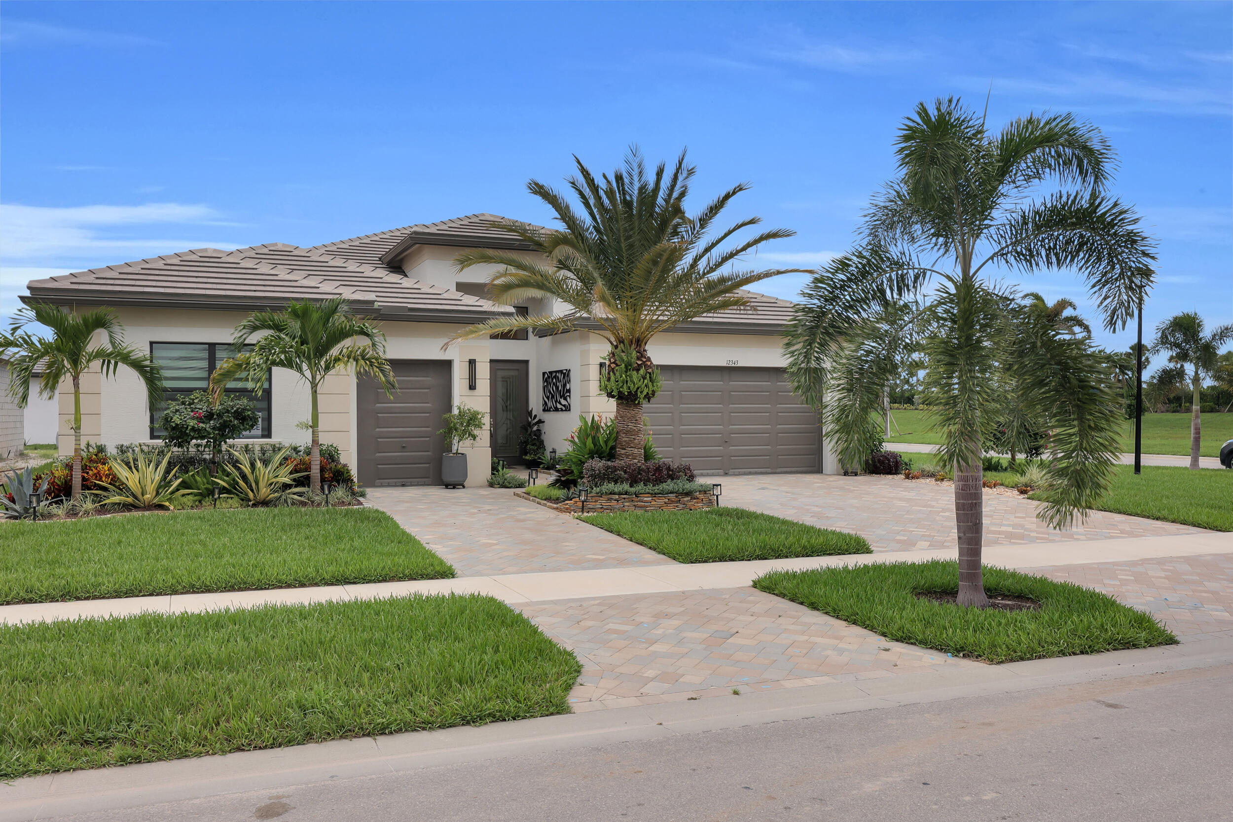 Contemporary stucco exterior with a tiled roof, paver driveway, two garage doors, and mature landscaping