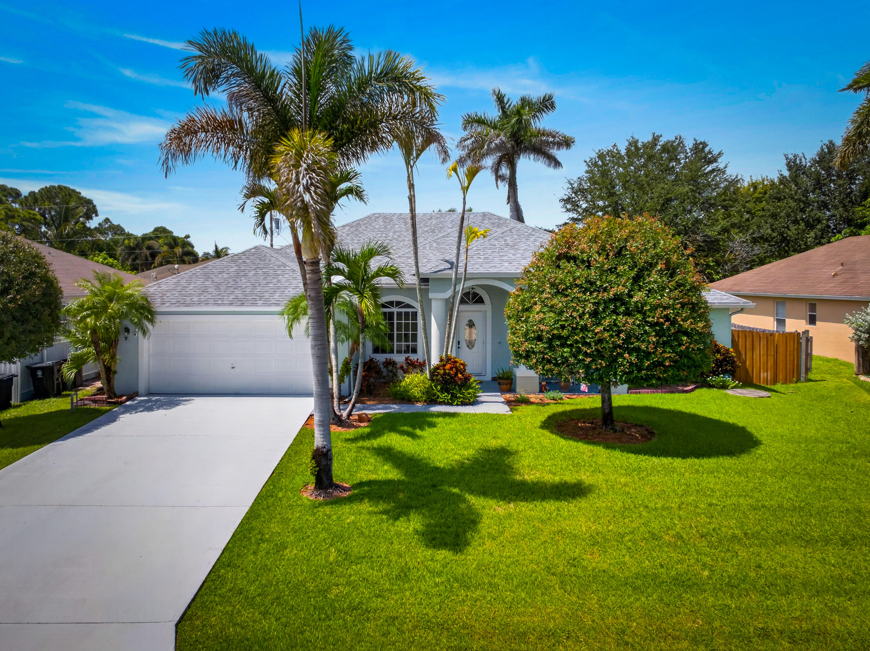 Light blue exterior with a white trim, mature palm trees, a well-kept lawn, an attached garage, and a concrete driveway