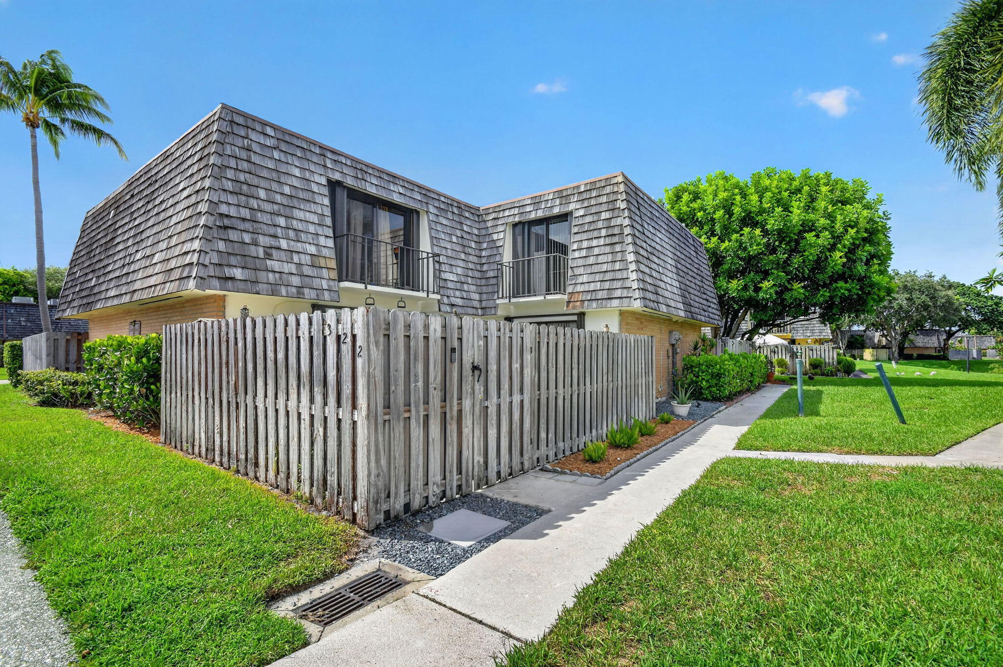Gambrel roofline with natural wood shingles, second-story balcony with iron railing, private fenced patio, mature landscaping, and an adjacent green space