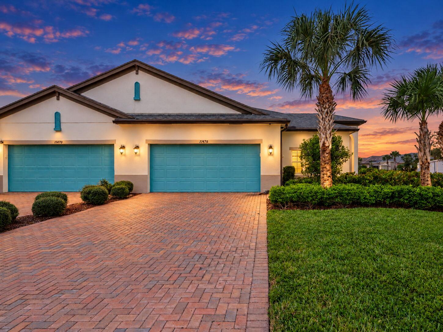 Stucco exterior with a tiled roof, paver driveway, two teal garage doors, and mature palm trees
