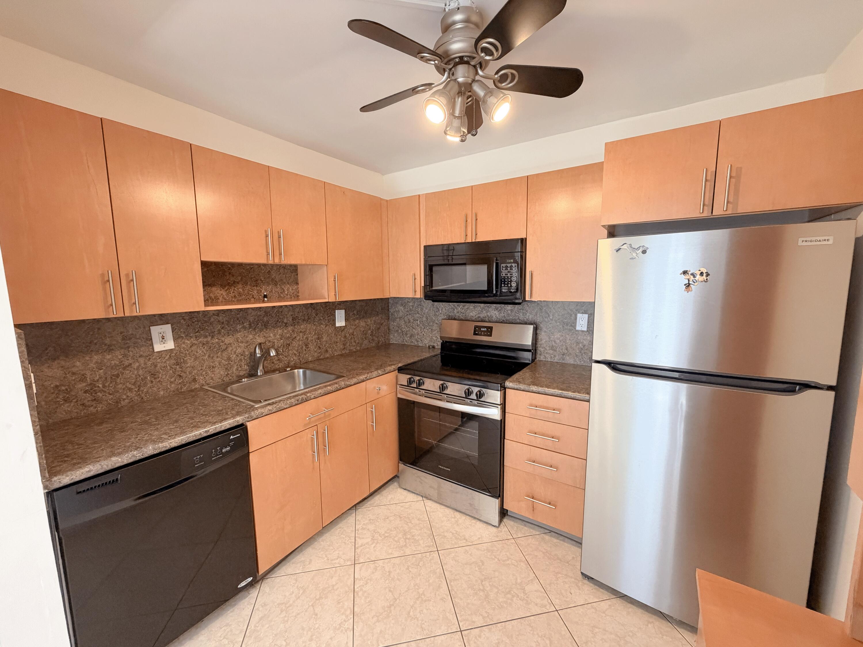 Kitchen featuring light wood-finish cabinetry, stone-look countertops, stainless steel refrigerator, black dishwasher, and a ceiling fan with integrated lighting
