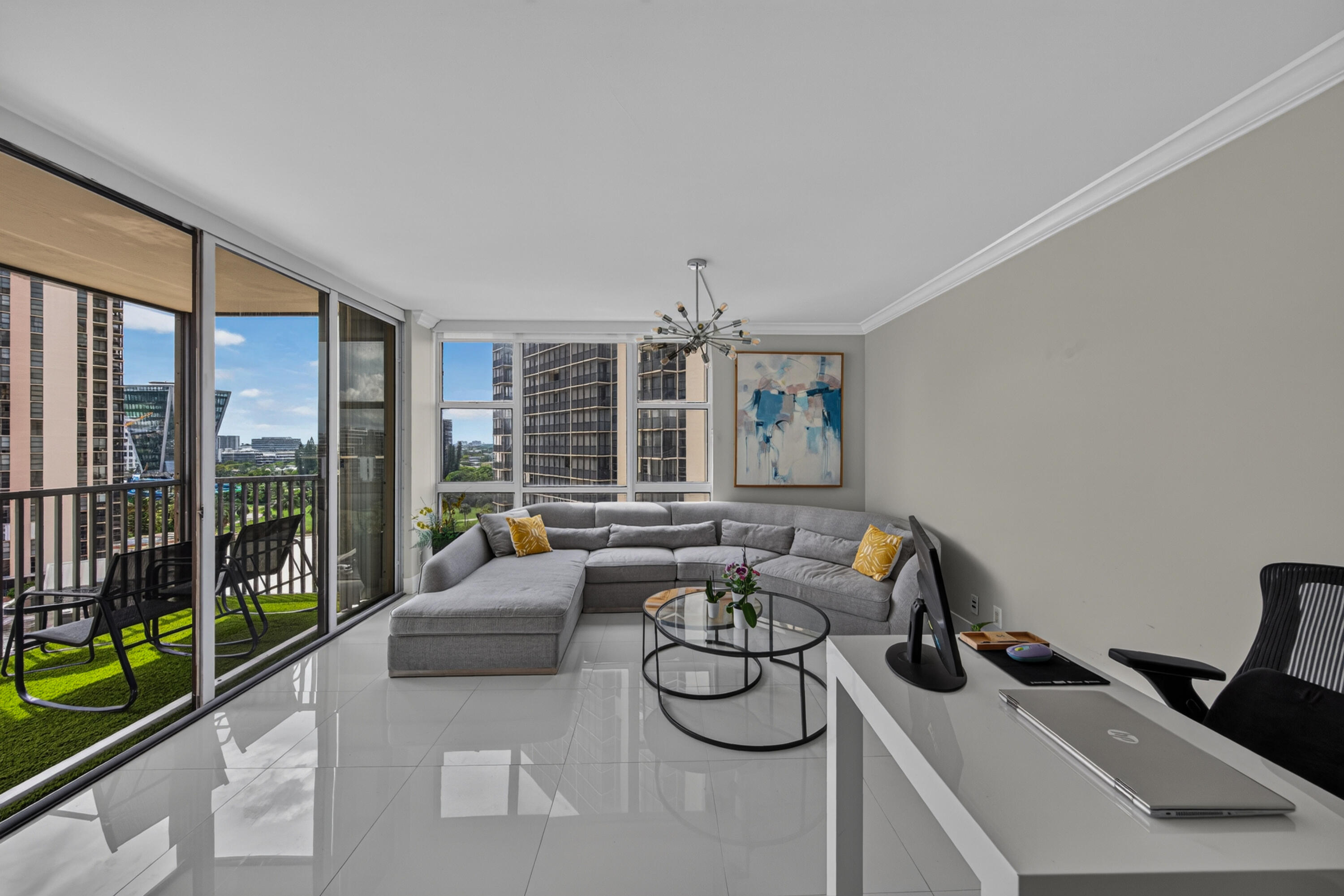 Contemporary living space featuring floor-to-ceiling glass doors opening to a private balcony with manicured lawn, polished light-finish flooring throughout, a gray upholstered sectional with chaise, round metal-framed coffee table, modern pendant chandelier, wall-mounted desk with integrated shelving, and expansive windows showcasing skyline and outdoor vistas