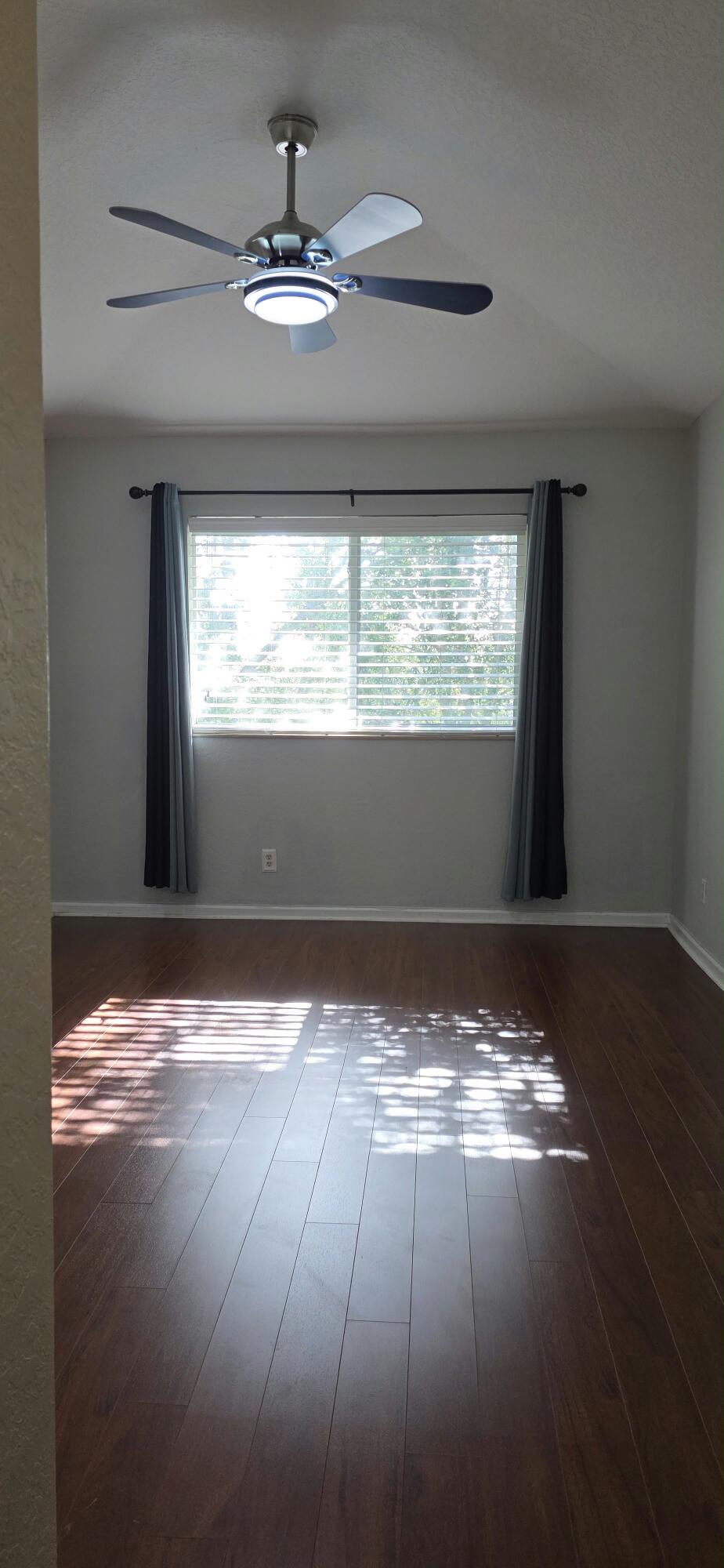 Wood-finish flooring with a satin sheen, large window with blinds, and ceiling-mounted fan with integrated lighting