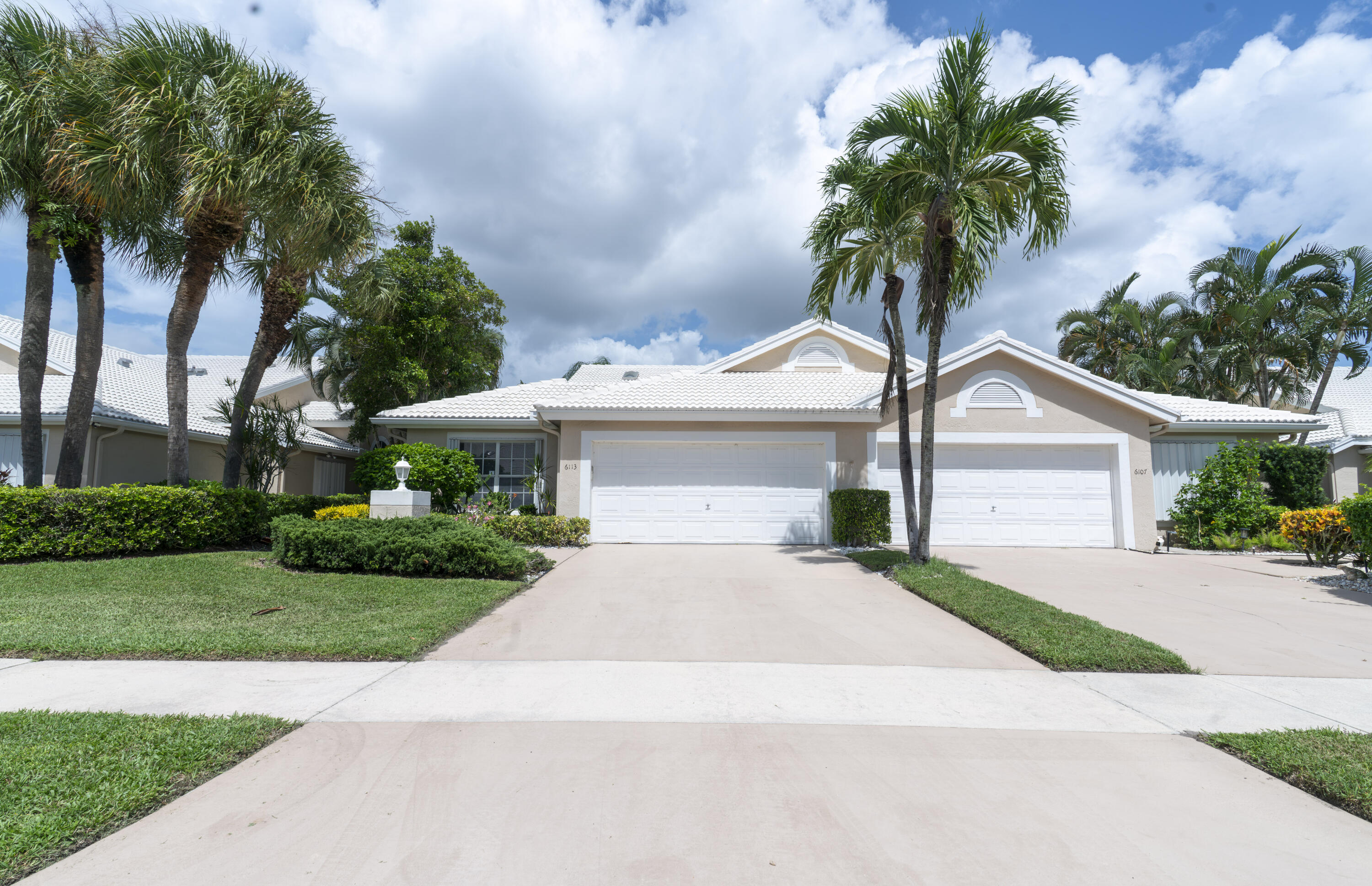 Exterior featuring a white tile roof, dual garage doors, a concrete driveway, established palm trees, and manicured landscaping