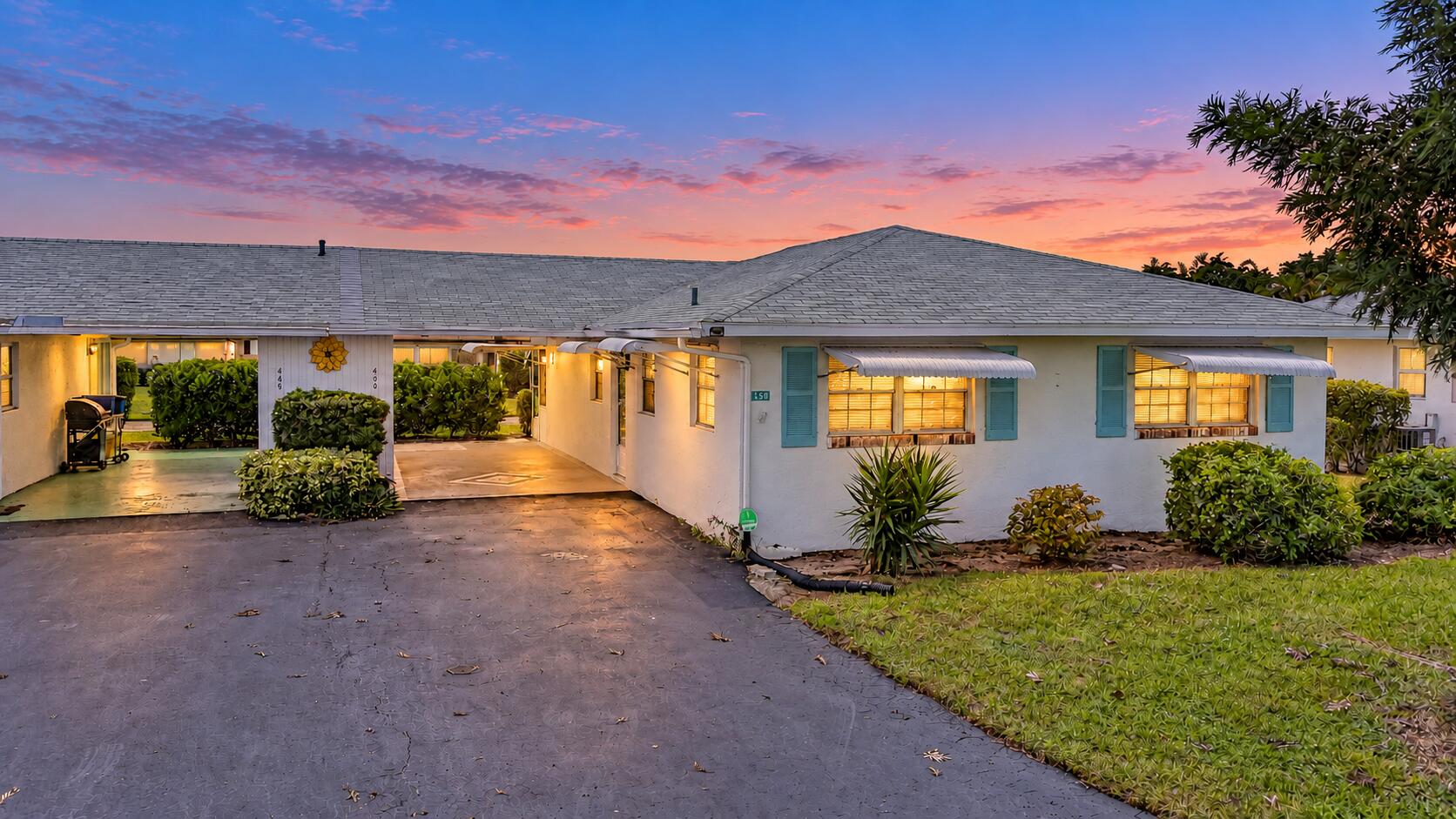 White exterior with light blue window shutters and awnings, asphalt driveway, covered carport, and established landscaping